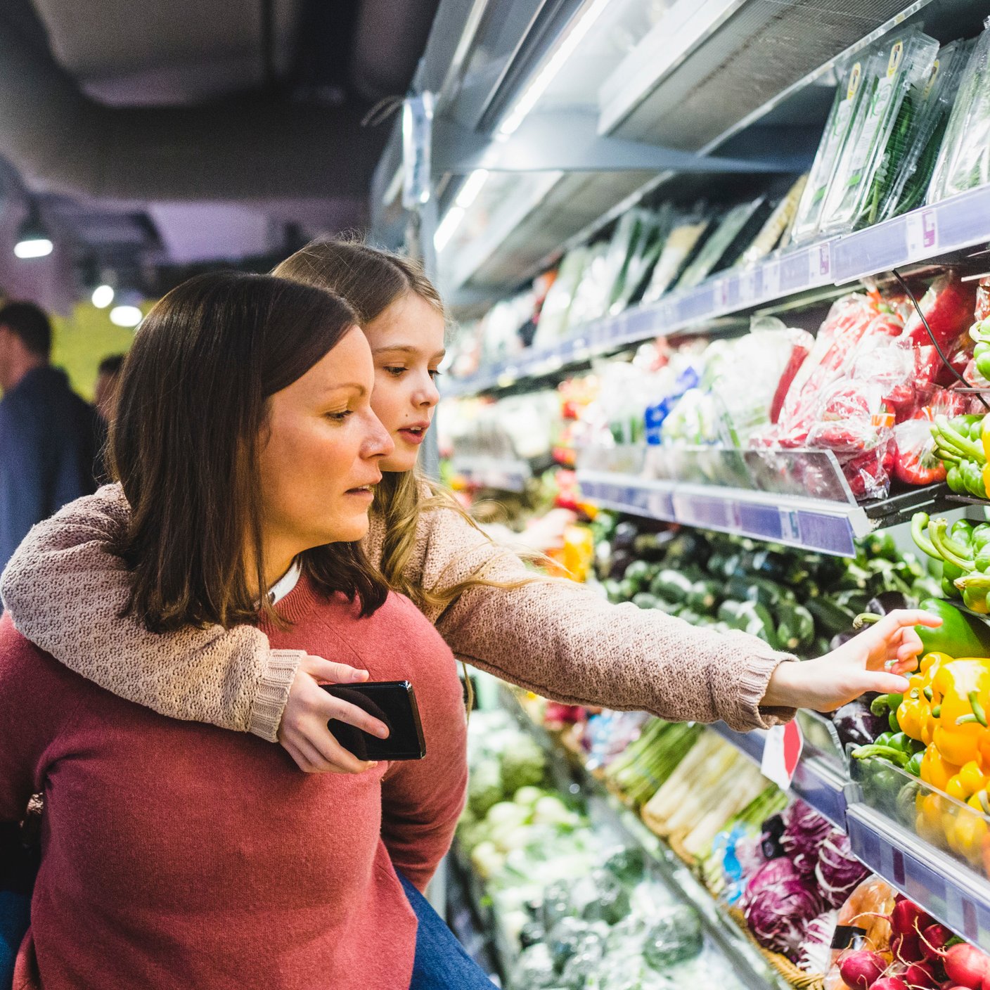 Cute daughter choosing bell peppers while being piggybacked by mother in store.