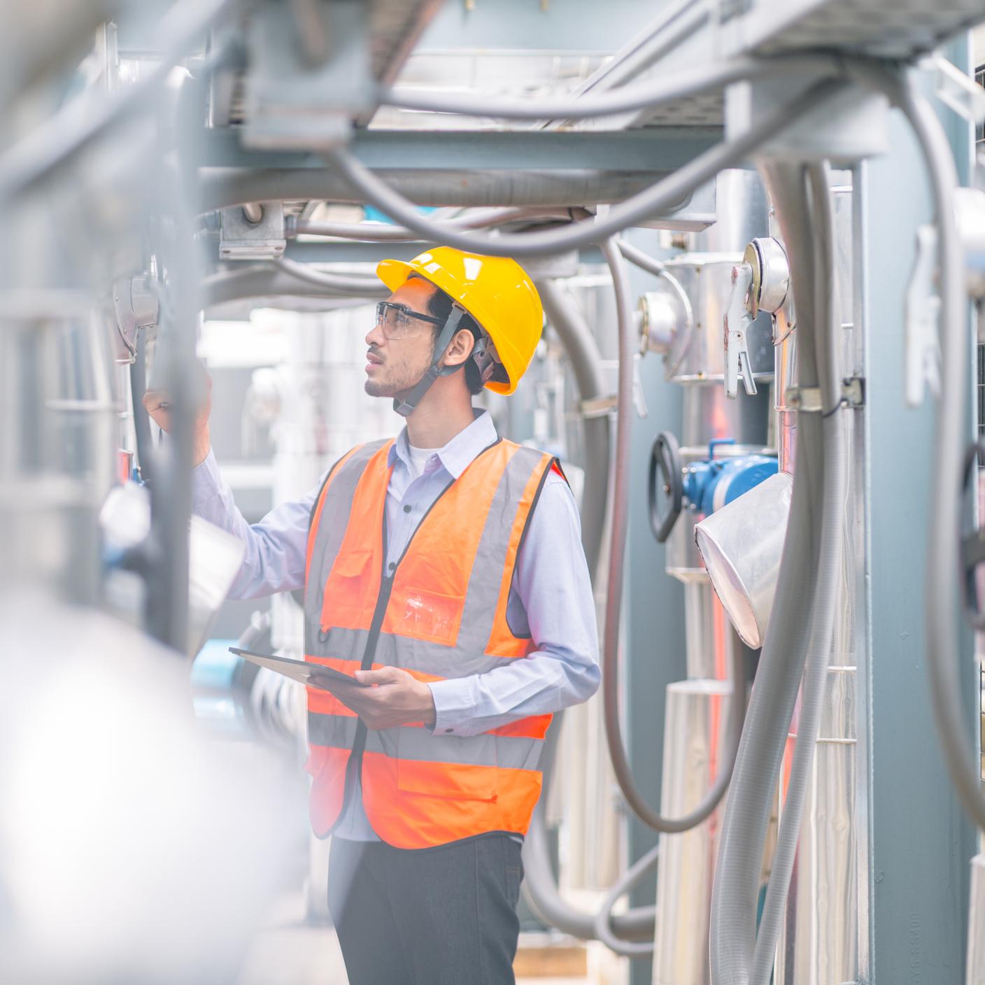 An engineer wearing a yellow hard hat and reflective vest is inspecting machinery with a clipboard in an industrial setting with visible piping.