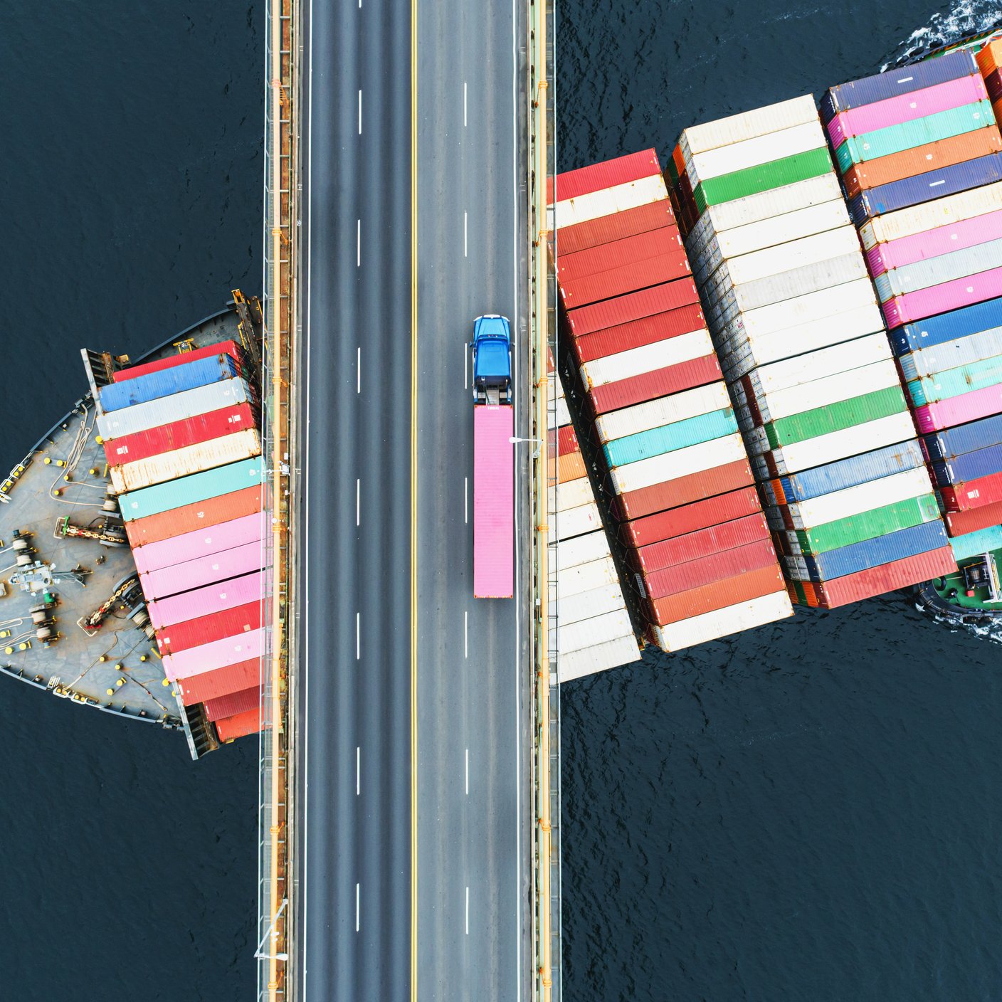 Vue aérienne d’un porte-conteneurs passant sous un pont suspendu. Camion semi-remorque avec un conteneur rose croisé au-dessus.