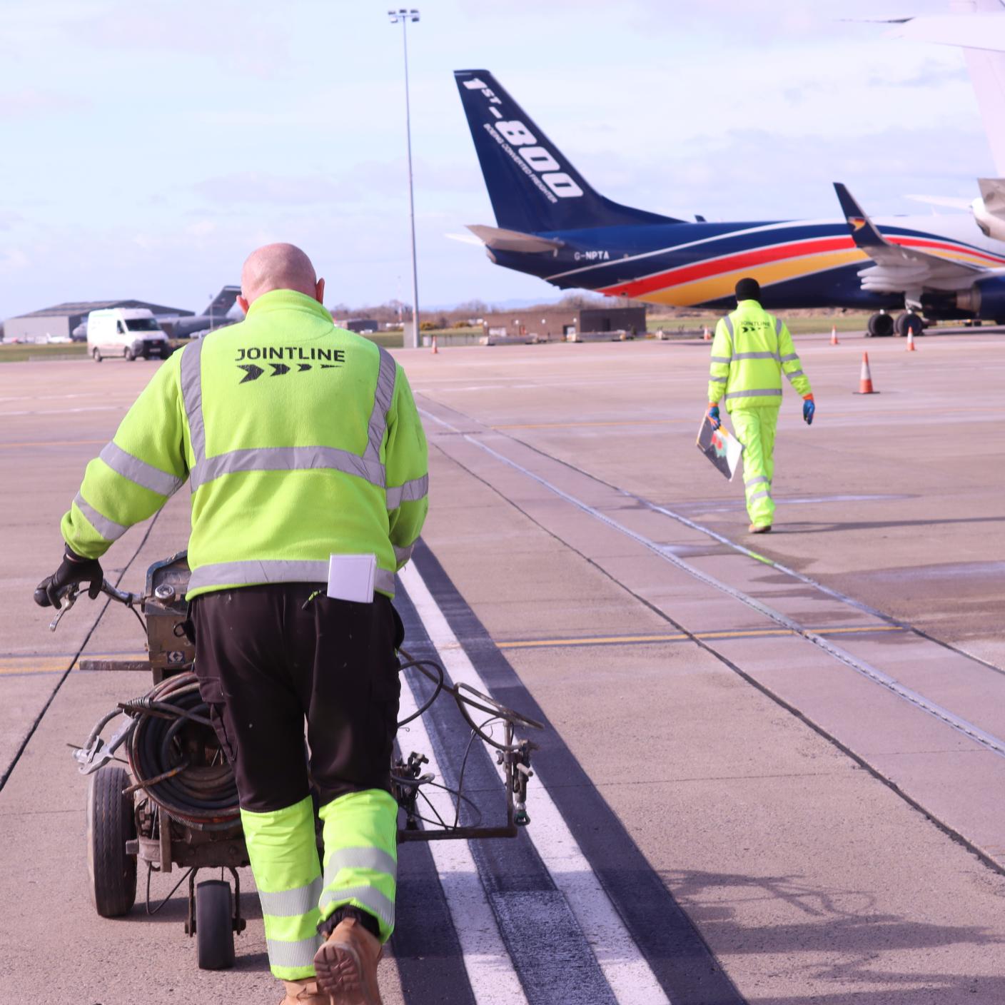 A worker in high‑visibility clothing pushes road‑marking equipment along an airport runway.
