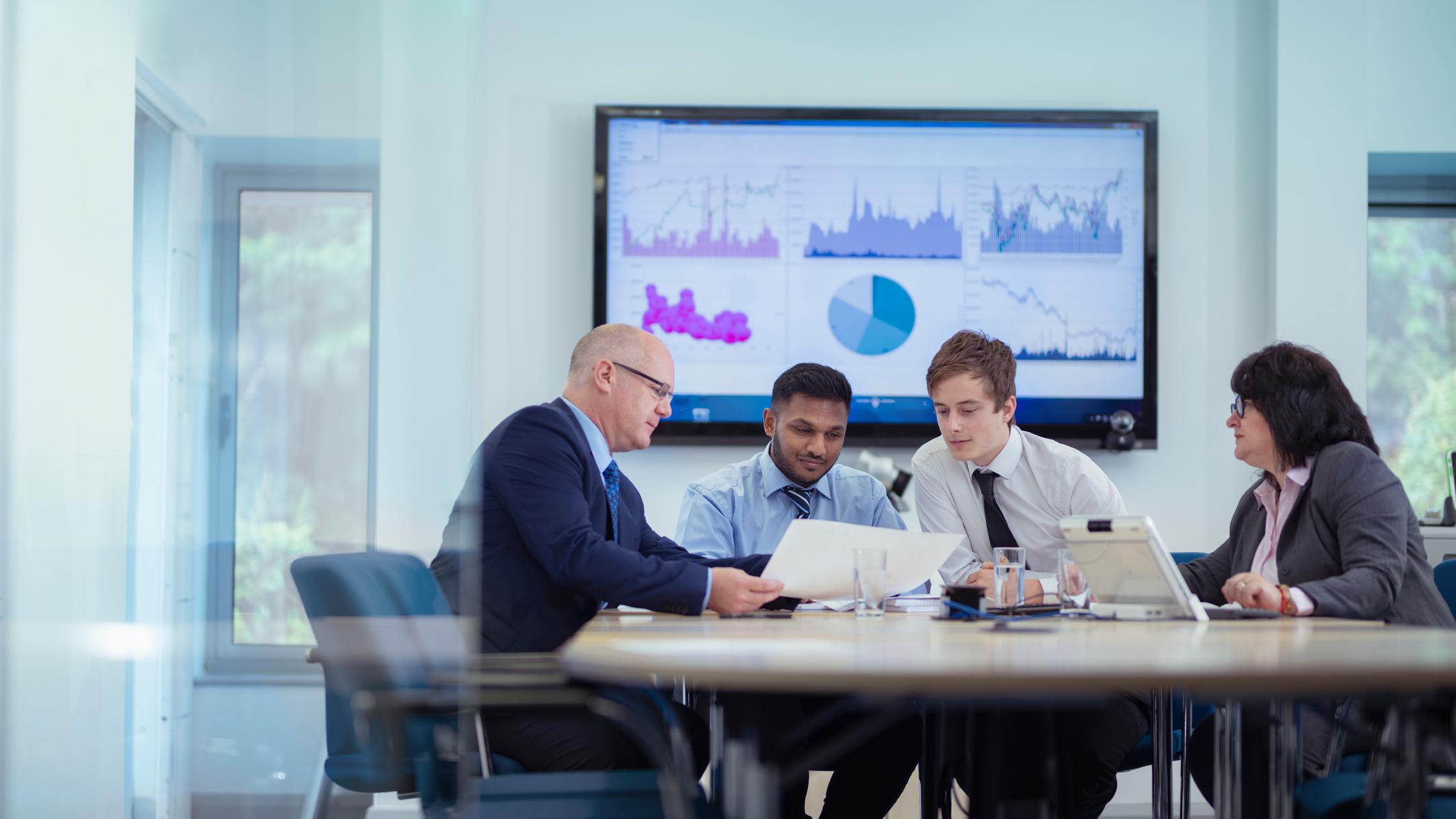 Multiple people in work office with electronic wall display in background.